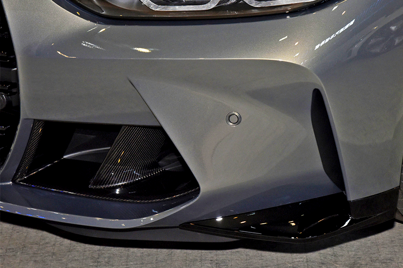 A sleek, silver car bumper features aerodynamic curves and a circular sensor, surrounded by glossy black and carbon fiber accents, resting on a smooth concrete floor.