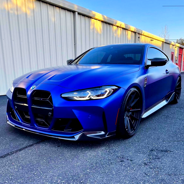 A blue sports car is parked on a paved area, with a large building featuring corrugated metal walls in the background under a clear blue sky.