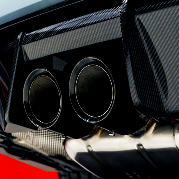 Two metallic exhaust pipes are positioned at the rear of a car, integrated within a carbon fiber diffusing panel. The background features a bold red surface.