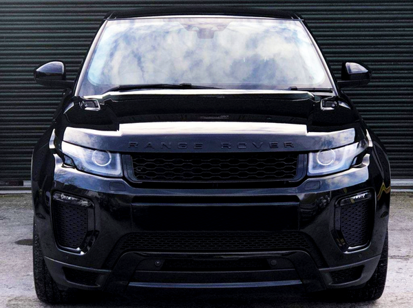 A black Range Rover stands stationary, facing forward, on concrete ground against a closed, corrugated metal door. The vehicle's headlights and grill are prominent. Text on the hood: "RANGE ROVER."