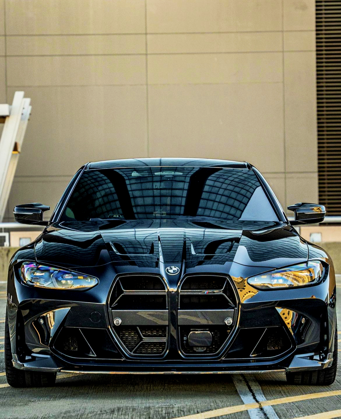 A sleek black car is stationary on a rooftop parking lot. Its modern design features aggressive front grilles and sharp headlights, with a reflective glass building visible in the background.