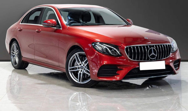 A red Mercedes-Benz sedan is parked indoors on a glossy floor, with bright lighting highlighting its sleek design and silver alloy wheels. The background is a plain dark wall.