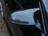 A carbon fiber car side mirror reflects its textured surface, attached to a sleek, black vehicle, with a gravel driveway visible nearby.