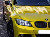 A yellow BMW car, parked, gleaming under sunlight on a cobblestone surface. The headlights and logo are prominently visible, surrounded by muted urban elements.