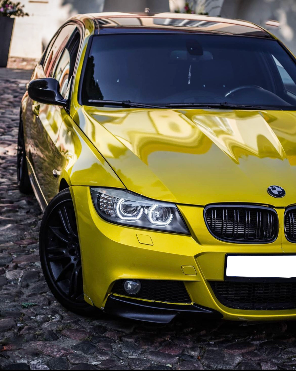 A yellow BMW car, parked, gleaming under sunlight on a cobblestone surface. The headlights and logo are prominently visible, surrounded by muted urban elements.
