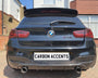 A black BMW M135i is parked on a driveway, featuring dual exhausts and a "CARBON ACCENTS" license plate. Surrounding houses display brick and white exteriors under a clear blue sky.