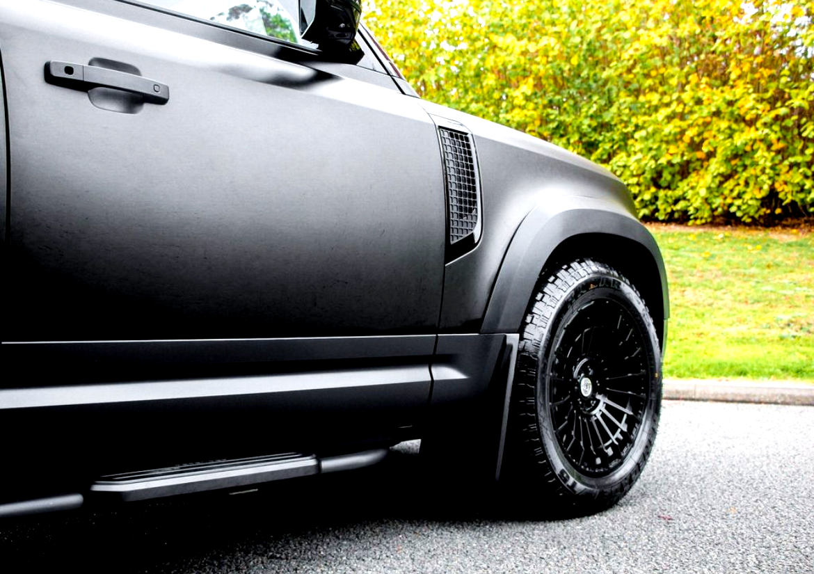 A black vehicle parked on a road features sleek body panels and large, textured tires. Trees with yellow-green foliage provide a natural backdrop.