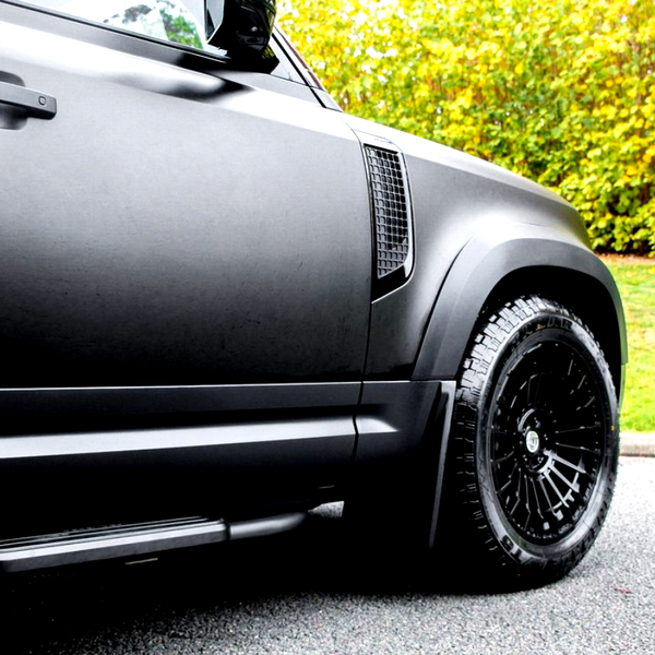 A black vehicle parked on a road features sleek body panels and large, textured tires. Trees with yellow-green foliage provide a natural backdrop.