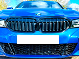 A blue car's front view showcases its black grille and logo, under a partly cloudy sky. License plate is blank, surrounded by trees and road surface visible below.