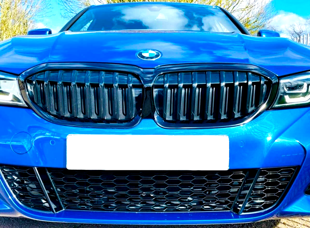 A blue car's front view showcases its black grille and logo, under a partly cloudy sky. License plate is blank, surrounded by trees and road surface visible below.