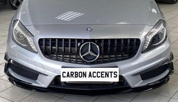 A silver car features a black grille with a prominent emblem, parked indoors on tiled flooring. The text on the license plate reads "CARBON ACCENTS."