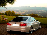 A silver BMW M4 is parked on cobblestones, surrounded by green fields and distant mountains under a clear sky with a tree and bench nearby.