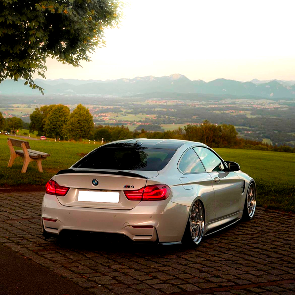 A silver BMW M4 is parked on cobblestones, surrounded by green fields and distant mountains under a clear sky with a tree and bench nearby.