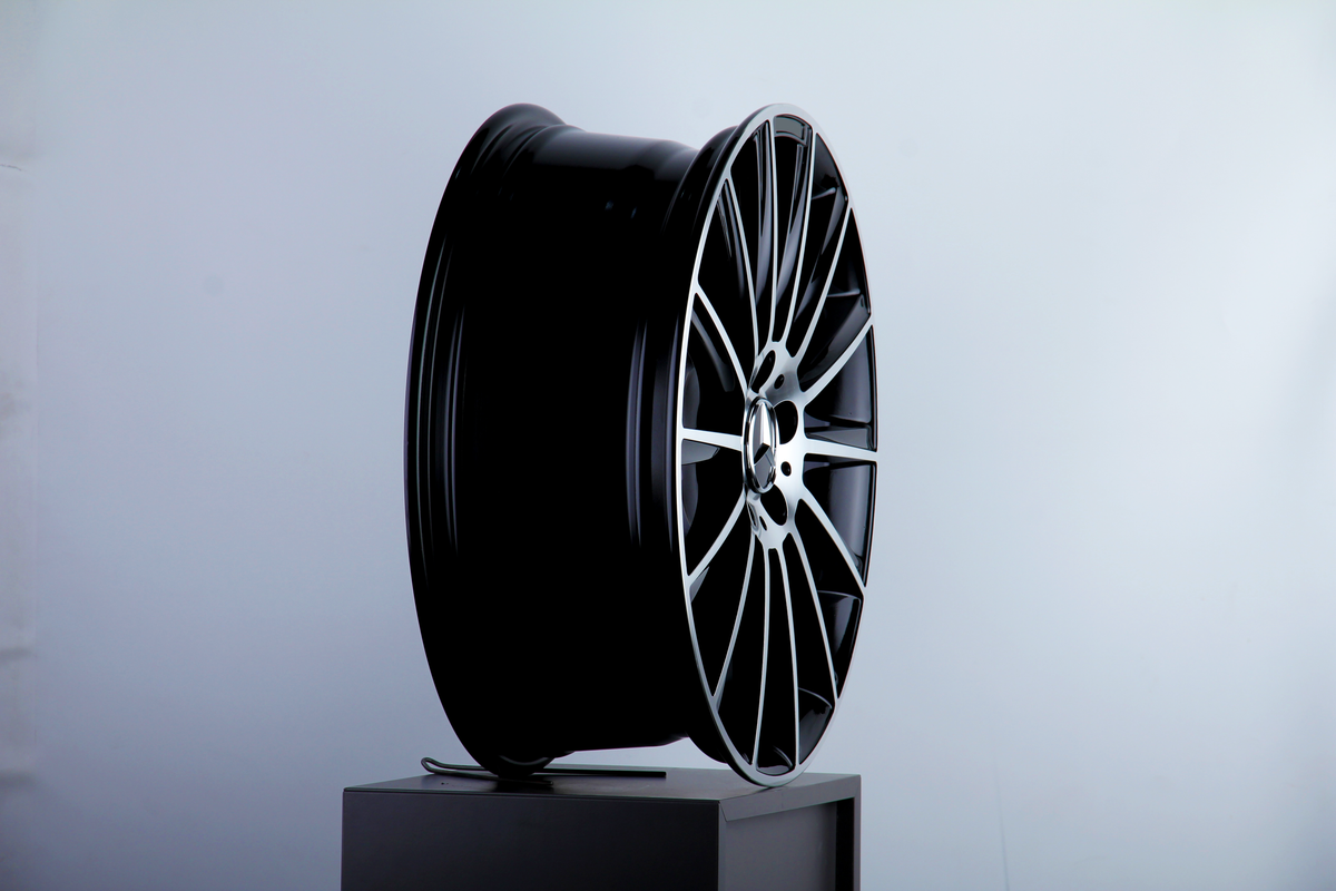 A sleek, black car wheel rim with intricate silver spokes sits upright on a gray pedestal in a minimalistic, light blue-gray setting.