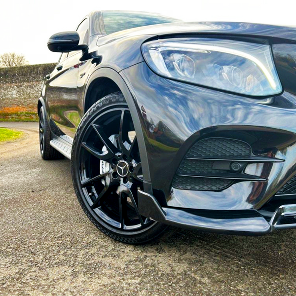 A sleek black car is parked on a paved area, featuring shiny alloy wheels and a modern headlamp design, with a stone wall and green grass in the background.