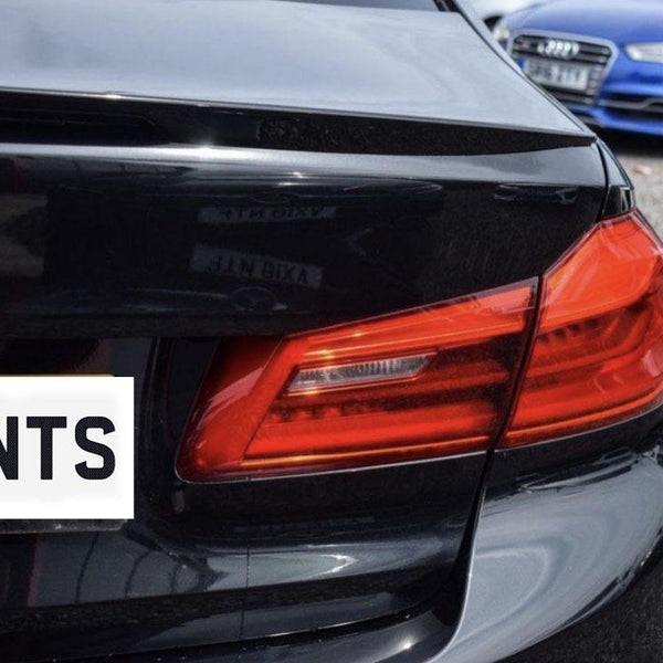 A black car with a red taillight is parked on a gravel surface. Text on the number plate reads "ENTS." A blue car is in the background.