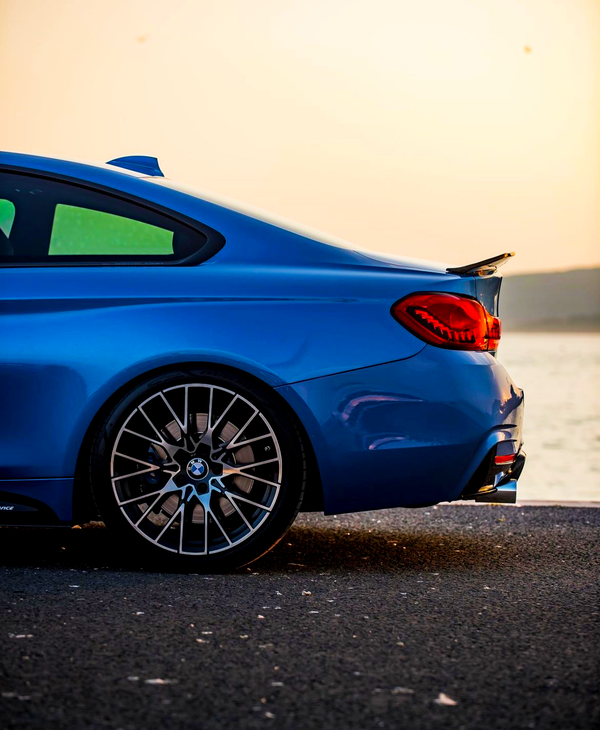 A blue sports car parked by a waterfront, showcasing its rear section with distinctive taillights and a prominent wheel in the foreground, against a serene sunset backdrop.
