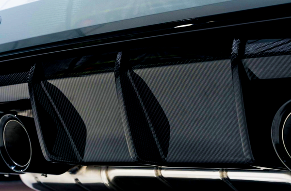 Carbon fiber rear diffuser with three vertical fins, flanked by metallic exhaust pipes, is mounted under a car's bumper in a well-lit garage setting.