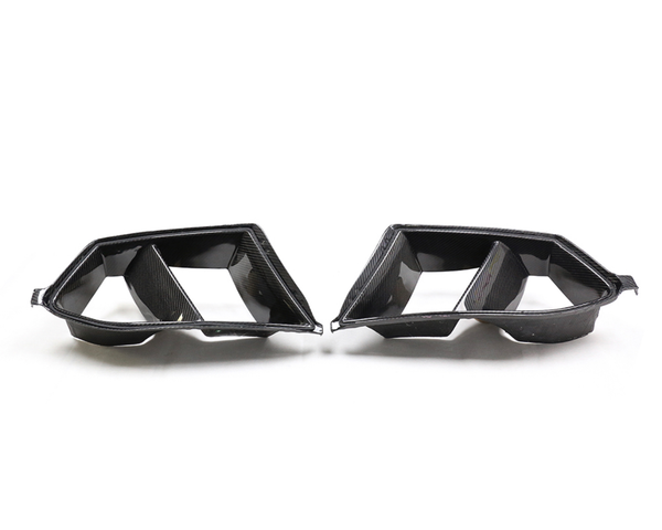 Carbon fiber exhaust tips, angular and dual-sectioned, lie side by side on a white background, reflecting light slightly from their glossy finish.