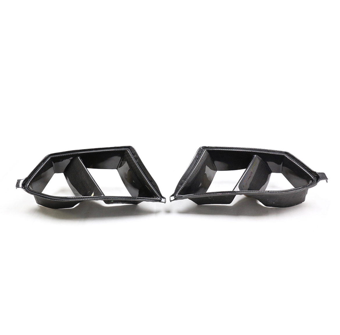 Carbon fiber exhaust tips, angular and dual-sectioned, lie side by side on a white background, reflecting light slightly from their glossy finish.