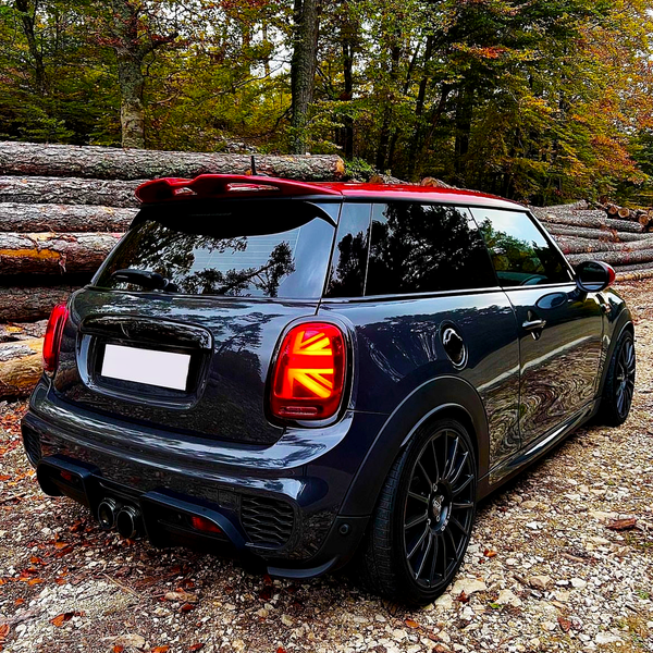 A dark-colored Mini Cooper with Union Jack tail lights is parked on a gravel path. Stacks of logs and autumn trees form the forested backdrop.