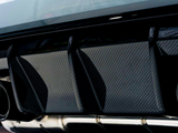 Dual exhaust pipes protrude from a sleek car rear diffuser with a carbon fiber texture, emphasizing performance and aerodynamics. The setting appears to be a modern garage or showroom.