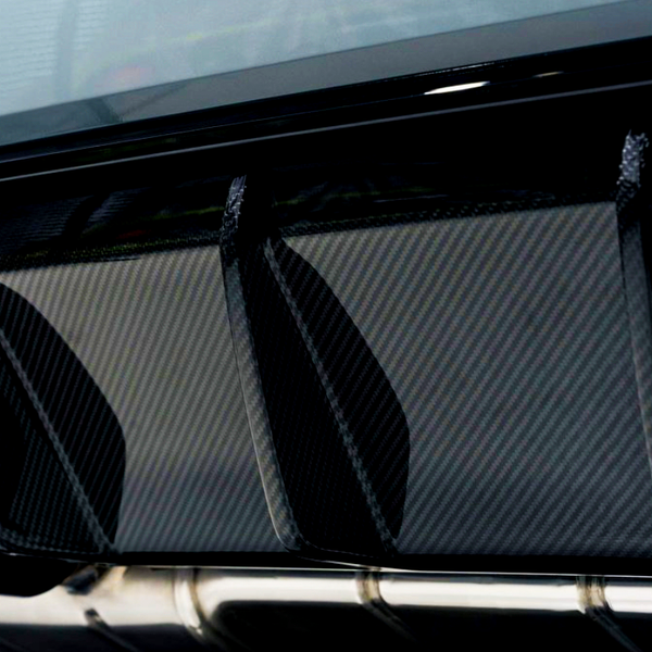 Dual exhaust pipes protrude from a sleek car rear diffuser with a carbon fiber texture, emphasizing performance and aerodynamics. The setting appears to be a modern garage or showroom.
