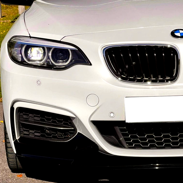 A white car's front showcases modern headlights and a signature grille. It's parked on asphalt, with grass and fallen leaves visible in the background.