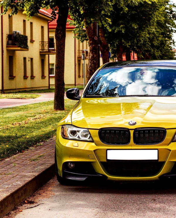 A bright yellow car is parked along a tree-lined street, adjacent to beige residential buildings with red roofs. The scene reflects a suburban or residential area.