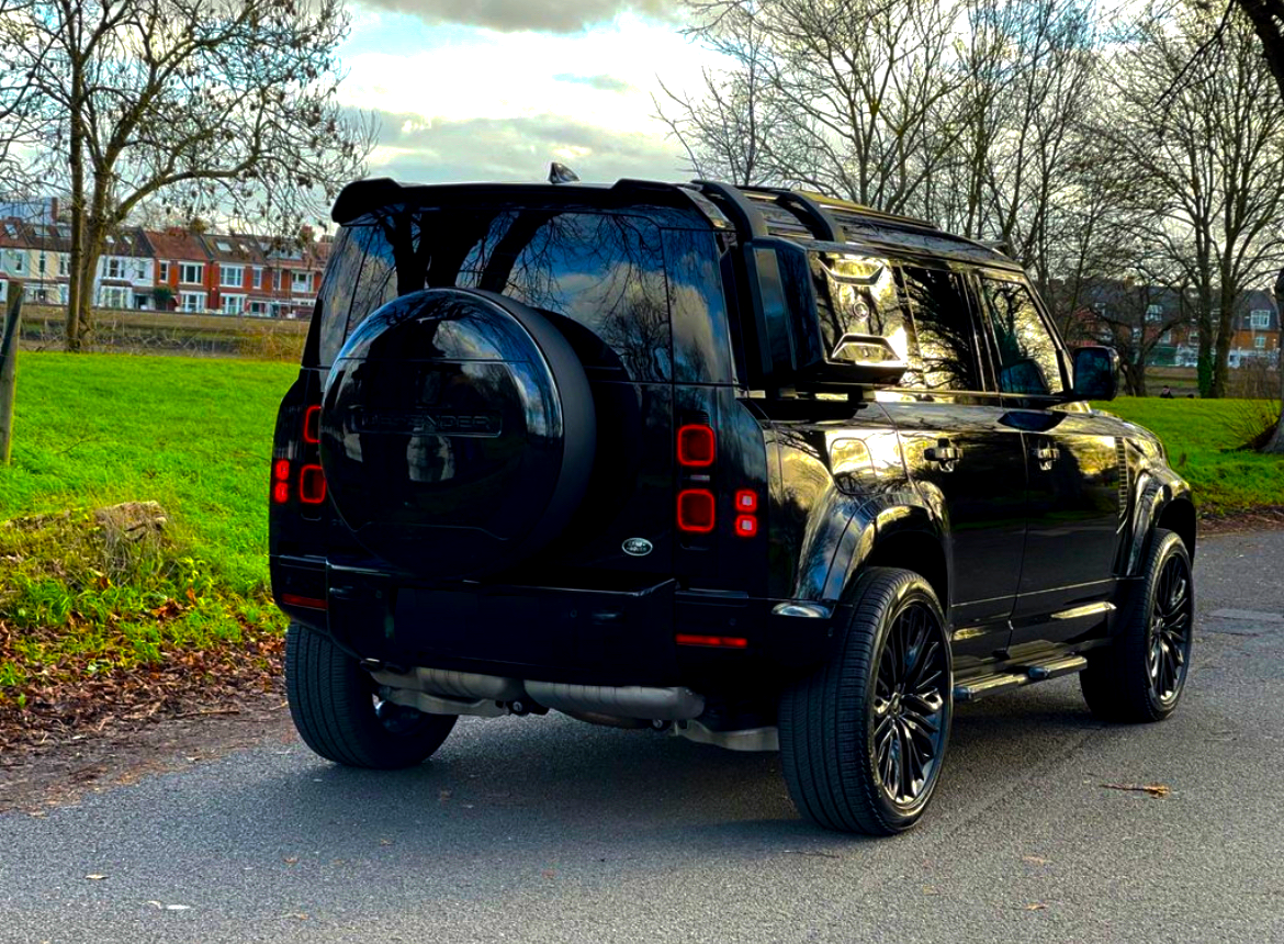 A black SUV is parked on a quiet road, with a grassy area and leafless trees surrounding it. Text "DEFENDER" is visible on the spare tire cover.