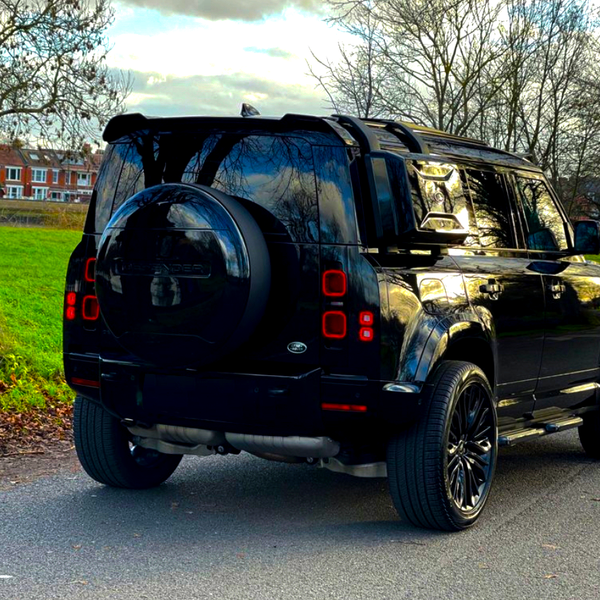 A black SUV is parked on a quiet road, with a grassy area and leafless trees surrounding it. Text "DEFENDER" is visible on the spare tire cover.