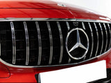 A chrome-accented grille with a prominent Mercedes-Benz emblem is centered on a red car's front, highlighted in a well-lit environment. It shows sleek vertical lines and a blank license plate.