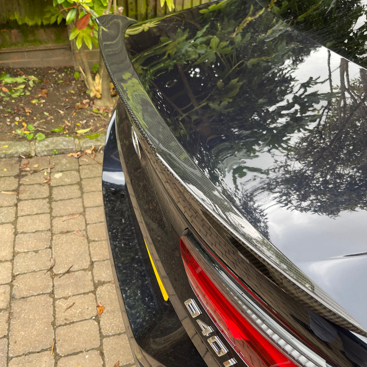 A car with a carbon fiber spoiler reflects foliage above, showing badge "640i" beside red tail lights, parked on a cobblestone driveway with a wooden fence and leaves nearby.