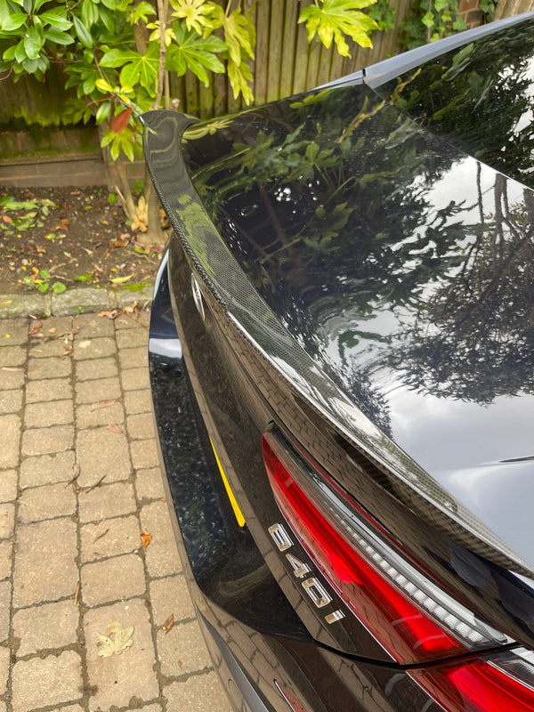 A car with a carbon fiber spoiler reflects foliage above, showing badge "640i" beside red tail lights, parked on a cobblestone driveway with a wooden fence and leaves nearby.