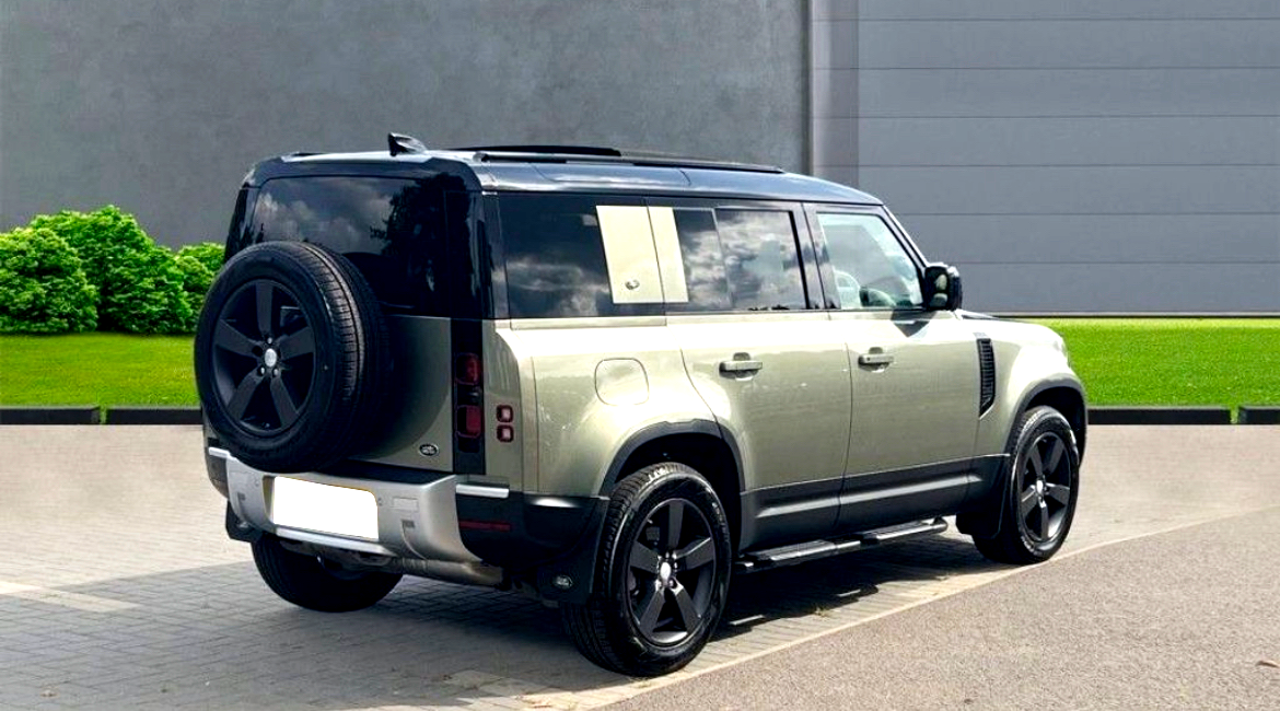 A green SUV is parked on a paved area with a spare tire mounted on its rear. It’s adjacent to a grassy lawn and a gray wall.