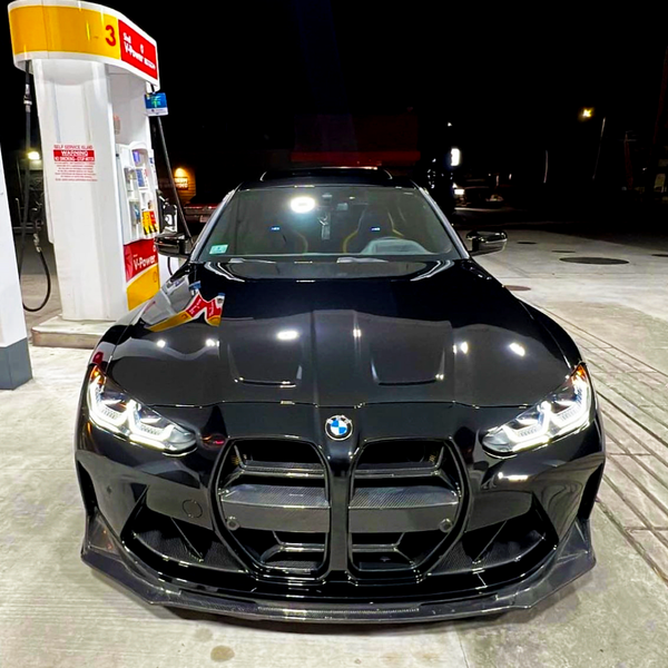 A black car, parked at a gas station at night, faces forward. It is under a Shell gas pump with visible signage that reads "Self Service Lane" and "V-Power."
