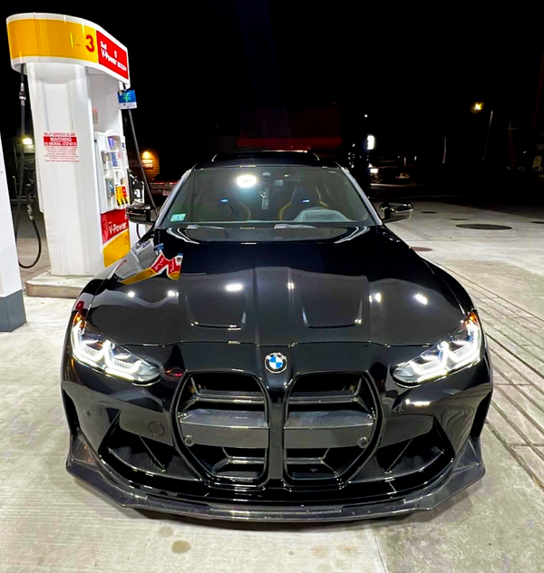 A black BMW car parked at a gas station pump. The setting is nighttime, with visible station lights and concrete ground. Pump labeled "3 Shell V-Power NiTRO+."
