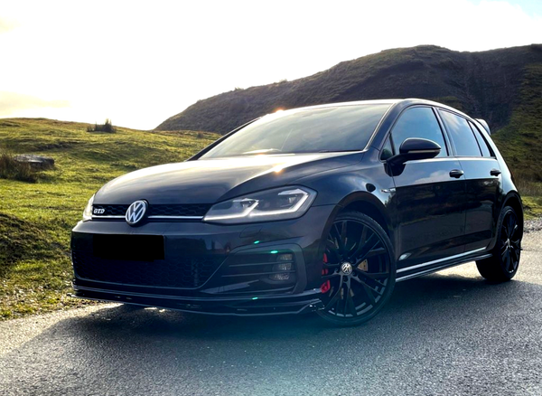 A black Volkswagen Golf GTD is parked on a paved road, with grassy hills and a bright, clear sky in the background.