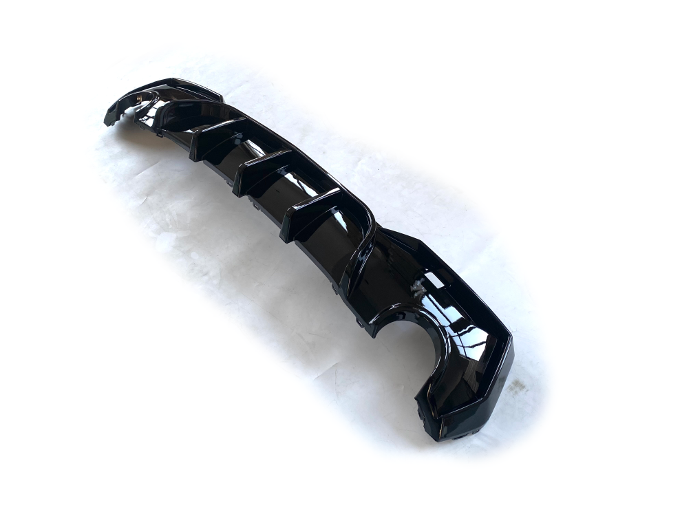 A glossy black car component, likely a diffuser, lies on a plain white surface. It features multiple aerodynamic fins and curves strategically positioned along its length.