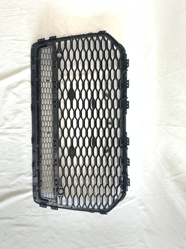 The black, hexagonal-patterned car grille is lying flat, showing its structured grid design on a white fabric backdrop, emphasizing its rectangular shape and multiple attachment clips.