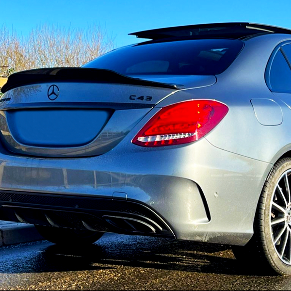 A silver Mercedes-Benz C43 car is parked on a wet road. The sunlight highlights its sleek design and red taillights, with trees and blue sky in the background.