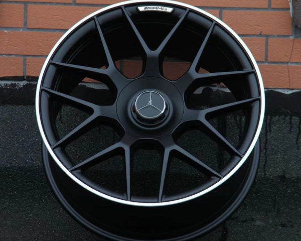 A black alloy wheel with a silver trim featuring a Mercedes-Benz logo rests against a brick wall. The AMG insignia is visible on the top part of the rim.