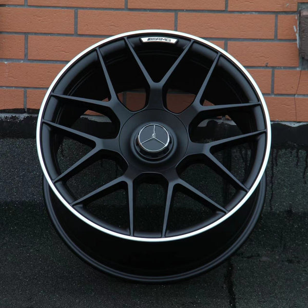 A black alloy wheel with a silver trim featuring a Mercedes-Benz logo rests against a brick wall. The AMG insignia is visible on the top part of the rim.