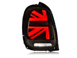 A black car taillight features illuminated red Union Jack design details, with a white horizontal light beneath, highlighting a modern automotive design against a plain background.