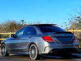 A gray Mercedes AMG car is parked on a road beside a wooden fence. The sky is clear, and tall shrubs line the background.