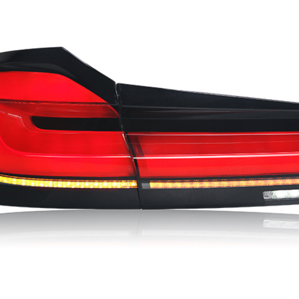 A car taillight, illuminated in red and amber, reflects on a white surface. The sleek design features multiple segments, with a glossy black frame accentuating its modern appearance.