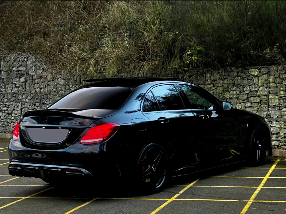 A black car is parked in a lot with yellow lines. It is positioned near a stone wall with overgrown greenery, suggesting a secluded or rural area.