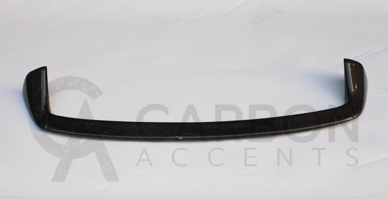 A carbon fiber car spoiler, curved and glossy, rests on a plain white surface. Text reads: "CARBON ACCENTS" with a partially visible stylized letter "A" behind the text.