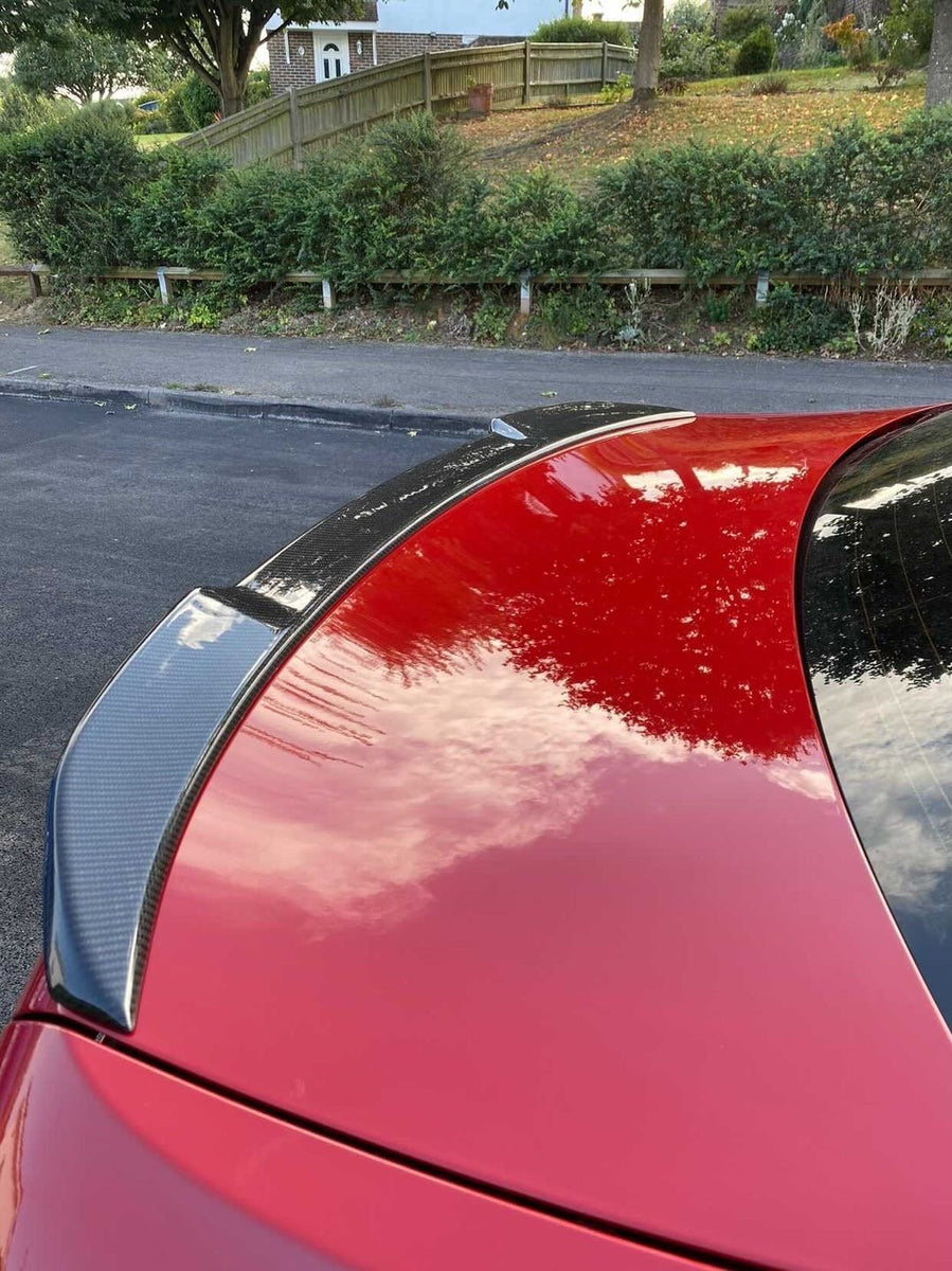 A red car's rear with a carbon fiber spoiler reflects clouds. It is parked on a street with lush greenery, bushes, and a wooden fence in the background.