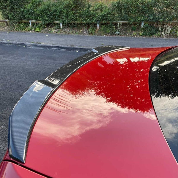 A red car's rear with a carbon fiber spoiler reflects clouds. It is parked on a street with lush greenery, bushes, and a wooden fence in the background.